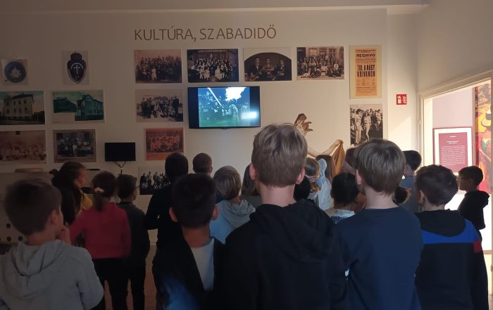 Egy múzeumi teremben gyerekek csoportja nézi a falra szerelt képernyőn megjelenő információkat. A képernyő körül régi fotók vannak elhelyezve.